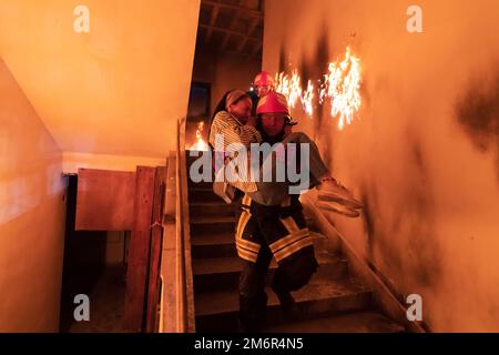 Der mutige Feuerwehrmann steigt die Treppe eines brennenden Gebäudes hinab und hält das errettete Mädchen in seinen Armen. Feuer eröffnen und ein Feuerwehrmann im Rücken Stockfoto