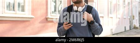 Ein Mann mit weißen Kopfhörern am Hals und einem Handy in der Hand, der die Straße in der Stadt entlang läuft. Stockfoto