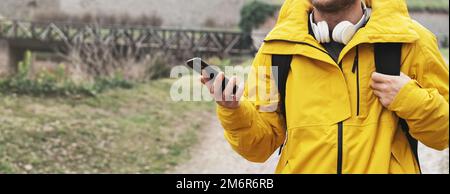 Ein Mann in einer gelben Reisejacke mit Kopfhörern am Hals und einem Smartphone in der Hand, der draußen unterwegs ist. Stockfoto