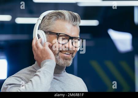 Nahaufnahme des Fotos eines erwachsenen, grauhaarigen Geschäftsmanns, eines grauhaarigen Mannes mit Kopfhörern, der sich Musikbücher und Podcasts online anhört, des Chefs am Arbeitsplatz im Büro. Stockfoto