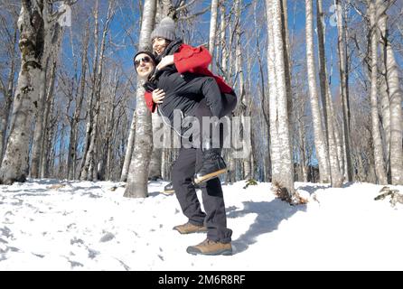 Glückliches junges Paar im Winterwald. Ein Mann, der Huckepack-Frauen-Ritt gibt. Mount Amiata, Toskana, Italien Stockfoto