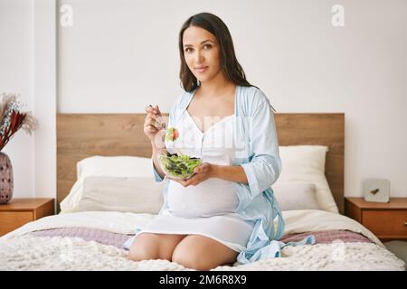 Porträt einer jungen schwangeren Frau im Pyjama, die im Bett einen frischen Salat isst Stockfoto