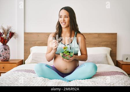 Porträt einer jungen Schwangeren in Sportbekleidung, die im Bett einen frischen Salat isst Stockfoto