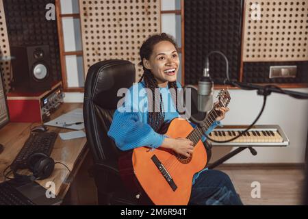 Fröhliche, talentierte Sängerin, die Gitarre spielt und in einem Musikstudio ein neues cooles Lied in ein Mikrofon singt Stockfoto