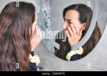 Eine attraktive, weiße, junge Frau, die in den Badezimmerspiegel schaut und ihre Falten um ihre Augen berührt Stockfoto