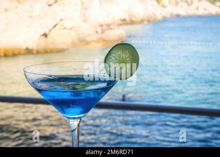 Ein blauer Cocktail in einem hohen Glas vor einem verschwommenen Hintergrund mit blauem Wasser am Strand Stockfoto