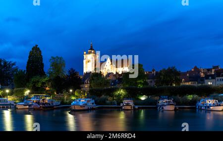 Nächtlicher Blick auf die beleuchtete katholische Kirche Notre Dame in Dole mit Hausbooten auf dem Doubs River im Vordergrund Stockfoto