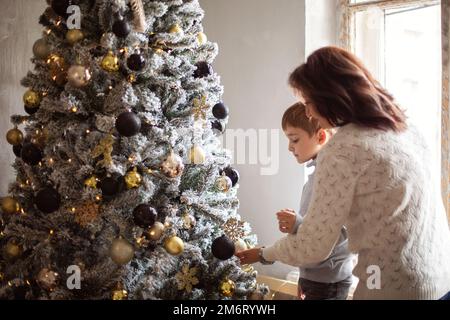 Ein kleiner Vorschuljunge, der Großmutter hilft, den Weihnachtsbaum zu Hause zu schmücken Stockfoto