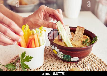 Frauenhände tauchen frische Gurken in hausgemachten Hummus mit Keksen, Petersilie und frischem Gemüse auf dem Tisch. Gesunde vegetarische rustikale Snacks aus der Nähe Stockfoto