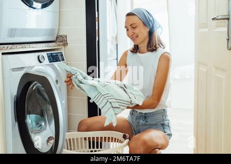 Eine Frau sitzt vor der Waschmaschine und lädt sie mit dreckiger Wäsche. Stockfoto