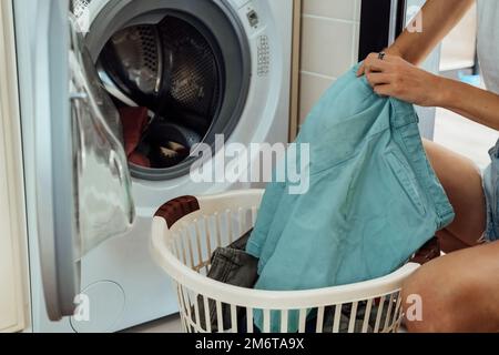 Eine Frau sitzt vor der Waschmaschine und lädt sie mit dreckiger Wäsche. Stockfoto