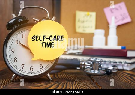 Medizinisches Konzept. Auf dem Wecker ein Aufkleber mit der Aufschrift "Self-care time". Im Hintergrund, unscharf, gibt es ein Stethoskop, ein Nasal Stockfoto