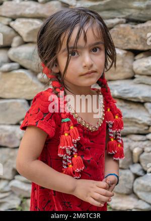 Yoged Village, Gorno-Badakshan, Tadschikistan - 08 21 2019 : Halbkörperporträt eines süßen kleinen Pamir-Mädchens in rotem Kleid mit traditionellen Haarnadeln Stockfoto