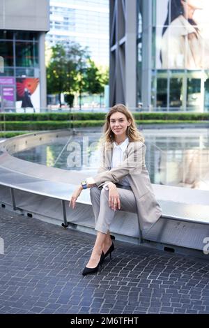 Vertikale Aufnahme einer erfolgreichen Geschäftsfrau im Anzug, die in der Nähe des Brunnens und der Geschäftshalle sitzt und selbstbewusst die Kamera anlächelt Stockfoto