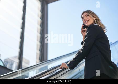 Porträt einer Geschäftsfrau in schwarzem Anzug, die auf die Rolltreppe steigt, mit dem Handy spricht. Eine Verkäuferin, die in der Stadt spaziert und eine ca. Stockfoto