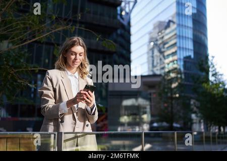 Junge Geschäftsfrau, die draußen auf der Straße steht. Büroangestellte, die ein Mobiltelefon verwenden und auf das Smartphone schauen Stockfoto