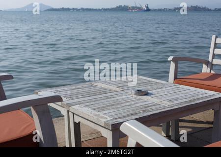 Ein Tisch in einem Freiluft-Restaurant mit Blick auf das Meer. Esstisch aus Holz und Holzstühle mit Kissen. Restaurant im Freien mit Meerblick Stockfoto