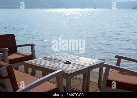 Ein Tisch in einem Open-Air-Restaurant mit Meerblick. Esstisch aus Holz mit Aschenbecher und Holzstühlen mit Kissen. Restaurant im Freien mit Meerblick Stockfoto