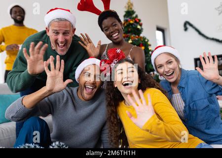 Bild von fröhlichen, vielfältigen Freunden, die zu Hause weihnachten feiern, Videoanrufe tätigen und winken Stockfoto