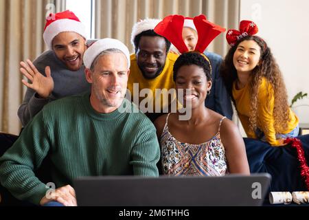 Bild von fröhlichen, vielfältigen Freunden, die zu Hause weihnachten feiern und Videoanrufe mit einem Laptop tätigen Stockfoto