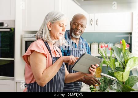 Glückliches, vielseitiges Paar, das Schürzen trägt und ein Tablet in der Küche benutzt Stockfoto