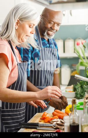 Glückliches, abwechslungsreiches Senior-Paar mit Schürzen und Kochen in der Küche Stockfoto