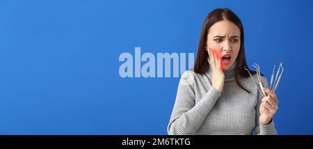 Ängstliche junge Frau mit zahnärztlichen Werkzeugen auf blauem Hintergrund mit Platz für Text. Nationaler Zahnschmerztag Stockfoto