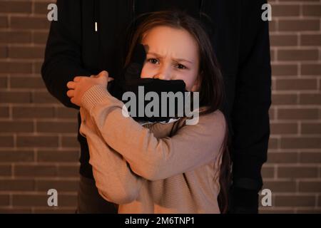 Terrorist und ängstliche kleine Geisel mit Backstein-Hintergrund Stockfoto