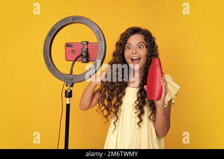 Teen girl, das vor der Kamera für Vlog auf gelbem Hintergrund isoliert spricht. Teenager arbeiten als Blogger halten Haarspülung oder Shampoo-Flasche Stockfoto