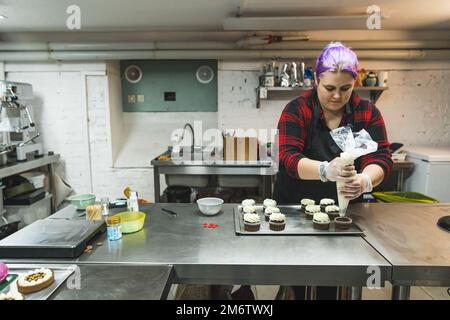 Baker bei der Arbeit. Fokussierte Frau, die eine Tüte in der Hand hält und Schokoladen-Muffins mit Glimmerpulver verz Bäckerei Küche. Hochwertiges Foto Stockfoto