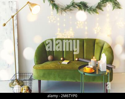 Grünes Velours-Sofa an weißer Wand mit Kranz und Weihnachtsbeleuchtung Stockfoto