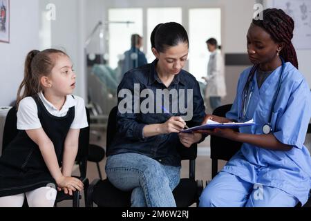 afroamerikanischer Arzt, der die Einreisepapiere für Kinder ausfüllt. Junge Frau, die eine kranke Tochter in eine medizinische Klinik eingeliefert hat. Schwester Oberärztin Mutter, kleines Mädchen in der Lobby des Sanatoriums. Stockfoto