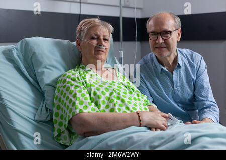 Ältere Frau nach einer Operation mit Bruder im Krankenhauszimmer. Eine kranke Frau wird in einer gerontologischen Klinik behandelt. Glücklicher alter Mann, der einen Verwandten im Sanatorium besucht. Stockfoto