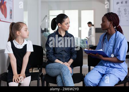 afroamerikanischer Arzt, der der jungen Mutter den Gesundheitszustand der Tochter erklärt. Erwachsene Frau, kleines Mädchen in der Sprechstunde im Kinderkrankenhaus. Kinderärztin und kleine Kinderschwester. Stockfoto