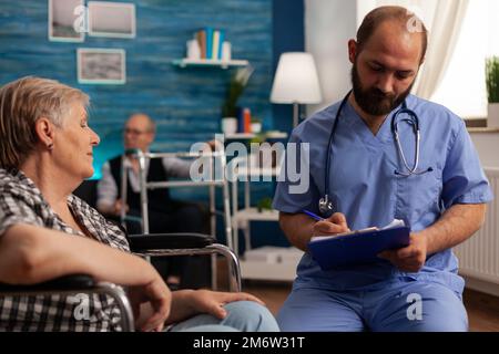 Männliche medizinische Assistentin trägt Uniform und Stethoskop ausfüllen Krankengeschichte des Seniorpatienten im Rollstuhl Notizen auf dem Klemmbrett. Frau, die der Krankenschwester persönliche Informationen übergibt. Stockfoto