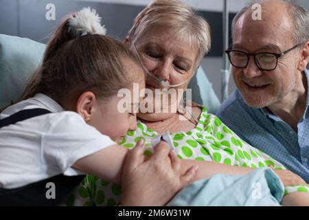 Älteres Ehepaar, das von einem kleinen Mädchen im Pflegeheim besucht wird. Lächelnde Enkelin zeigt Liebe zu glücklichen Großeltern, umarmt sich in der Klinik. Nahaufnahme der Umarmung zwischen Kleinkind, alte Dame. Stockfoto