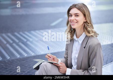 Geschäftsfrau im Anzug, schreibt smth, arbeitet an Ideen im Notizbuch, sitzt draußen im Stadtzentrum Stockfoto