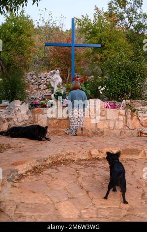 Eine Frau, die am Blauen Kreuz in Medjugorje kniet und betet. Die abgebildeten freilaufenden Hunde sind ein üblicher Anblick. Stockfoto