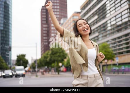 Porträt einer glücklichen asiatischen Frau, tanzen und Freude fühlen, triumphieren, die Hand in Siegesgeste hochheben, auf der Straße feiern Stockfoto