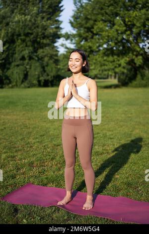 Vertikale Aufnahme eines lächelnden asiatischen Mädchens, das Yoga macht, Stretching-Übungen im Park macht, auf einer Gummimatte an der frischen Luft steht Stockfoto