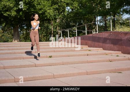 Gesundes Fitness-Mädchen, das draußen auf der Straße läuft, Uniform trägt, an der frischen Luft joggt und Musik mit kabellosen Kopfhörern hört Stockfoto