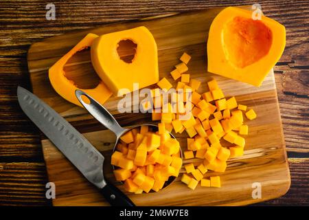 Butternusskürbis, der auf einem Holzschneidebrett in kleine Würfel geschnitten wurde: Gewürfelter Winterkürbis auf Holzhintergrund mit einem Küchenmesser Stockfoto