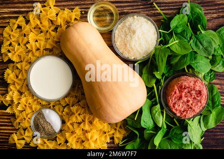 Cremig geröstete Butternusskürbis-Pasta mit Wurst und Spinat Zutaten: Farfalle-Pasta, Winterkürbis und andere rohe Zutaten auf einem Holztisch Stockfoto