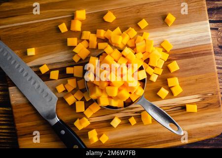 Butternusskürbis, der auf einem Holzschneidebrett in kleine Würfel geschnitten wurde: Gewürfelter Winterkürbis auf Holzhintergrund mit einem Küchenmesser Stockfoto