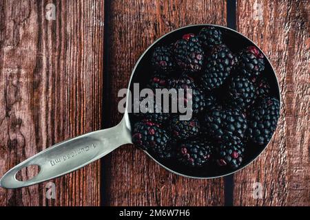 Frische Brombeeren in einem Messbecher auf einem Holztisch: Ganze Beeren in einem Messbecher aus Edelstahl auf rustikalem Holzhintergrund Stockfoto