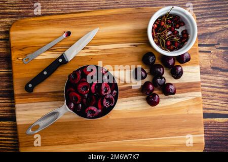 Halbierte und entsteinte Kirschen auf einem Holzschneidebrett: Rohe Kirschen mit Werkzeugen zum Schneiden und Gruben von oben gesehen Stockfoto