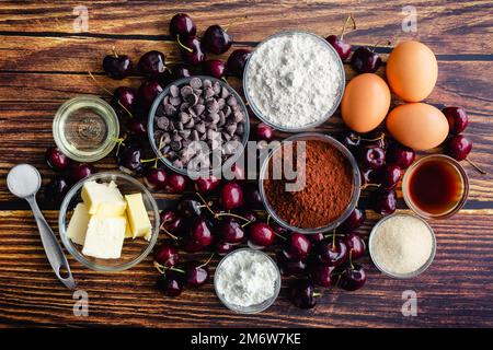 Roasted Chocolate Cherry Brownie Zutaten auf einem rustikalen Holztisch: Frische Kirschen, Kakaopulver, Schokoladenchips und andere rohe Zutaten Stockfoto