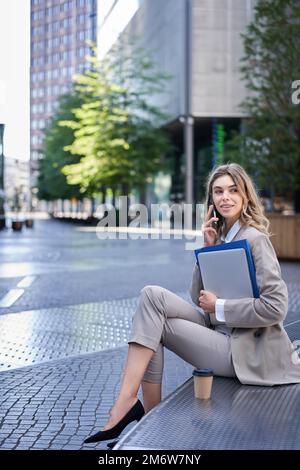 Vertikale Aufnahme einer jungen Frau, die in der Nähe eines Bürogebäudes sitzt, Laptop und Ordner mit Arbeitsdokumenten in der Hand hält, jemanden auf Mo anruft Stockfoto