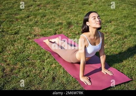 Lächelnde junge Sportlerin streckt sich im Park auf einer Gummimatte, macht Yoga-Asana-Übungen, trainiert an der frischen Luft in Fitnesskleidung Stockfoto