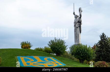 Die Statue des Vaterlands vor dem blauen Himmel. Ein Wappen der Ukraine auf dem Rasen, mit blauen und gelben Steinen gesäumt. Die Tride Stockfoto